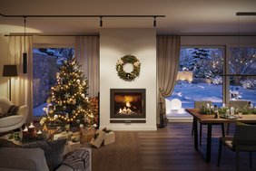 Im festlich geschmückten Wohnzimmer steht ein beleuchteter Weihnachtsbaum mit Lichterketten und Kugeln in dezenten Farben (Weiß, Gold, Silber)neben einem Kamin, über dem ein Kranz hängt. Vor dem Baum liegen verpackte Geschenke. Rechts befindet sich ein Esstisch, der mit Tannenzweigen und Kerzen dekoriert ist. Große Fenster geben den Blick auf einen verschneiten Garten frei, in dem leuchtende Dekorationsobjekte platziert sind.