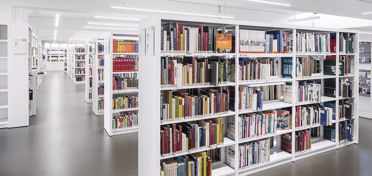 In einer Bibliothek sind abgependelte Deckenleuchten entlang der Laufwege montiert sowie parallel zu den hintereinander angeordneten Regalreihen. Die Titel der Bücher sind gut zu erkennen.   