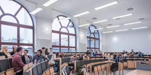 Große bodentiefe Fenster lassen viel Tageslicht in den Höhrsaal. Längs zu ihnen sind Pendelleuchten montiert, die eine blend- und schattenfreie Atmospäre schaffen. Wandleuchten ergänzen die Allgemeinbeleuchtung.