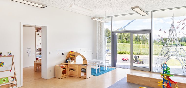 Zu sehen ist ein Spielzimmer in einer Kindertagesstätte. Bodentiefe Fenster lassen viel Tageslicht hinein und Pendeleuchten an der Decke steuern künstliches Licht hinzu.