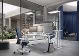 Das modern eingerichtete Büro hat einem Schreibtisch mit zwei Bildschirmen und einer Akustik-Tischtrennwand zum gegenüberliegenden Arbeitsplatz. Rechts und links je eine Stehleuchte, die beide Tische ausleuchtet. 