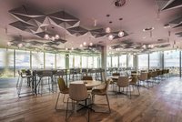 Eine moderne Kantine mit zwei verglasten Gebäudeseiten wird vom farbigen Licht runder Glaspendelleuchten erhellt.