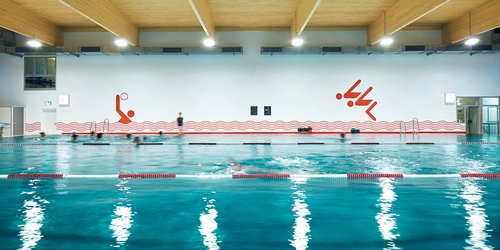 Indoor Schwimmbäder Blick in eine große Schwimmhalle mit Kuppeldecke und Sitztreppen an den Längsseiten des Beckens: Gezeigt wird der Schwimmbetrieb am Abend, wenn alle Lichter eingeschaltet sind: ein durchgehendes Lichtband am unteren Bereich der Kuppel und am oberen Rand der Wände.