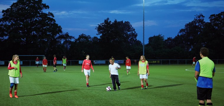 Eine gemischte Mannschaft trainiert Fussball auf einem Rasenplatz, der von Flutlichtern erhellt wird.
