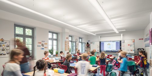 Ein großes längliches Klassenzimmer mit parallel zum Fenster angebrachten Pendelleuchten, die von der Tafel bis an die gegenüberliegende Wand reichen und viel Helligkeit geben.