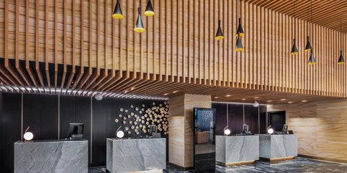 Eine luxuriöse Hotelrezeption ist mit dunklen Marmorböden und Holzwänden gestaltet. Extravagante trichterförmige Pendelleuchten hängen von der Decke, auf den Empfangstresen stehen kugelförmige Tischleuchten.