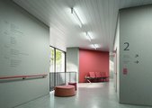 Eine blendfreie und gleichmäßige Ausleuchtung verhindert störende Schatten im Flur, die zu Stürzen führen können. (Foto: licht.de/Waldmann) Ein Klinikkorridor mit einer Sitzbank zum Warten und Richtungshinweisen an den Wänden. Durch ein großes Fenster scheint Tageslicht und drei Deckenleuchten geben künstliche Beleuchtung.